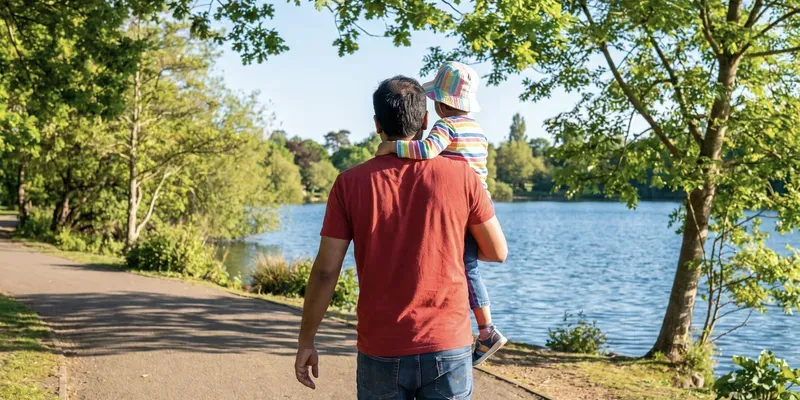 A father, seen from behind, walks a lakeside path carrying his child on his hip.