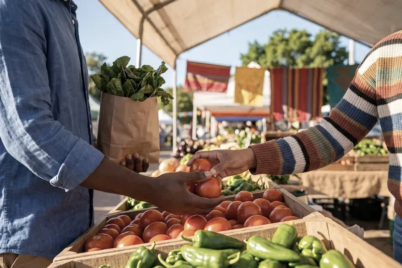 Hands choose tomatoes and peppers at a sunny market stall.